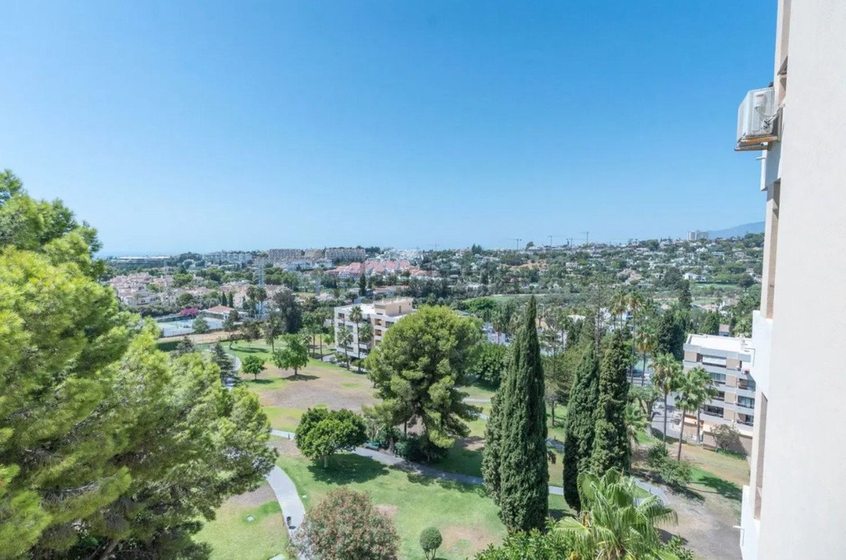 View from a balcony overlooking a green park area with trees and a cityscape in the distance under a clear blue sky.
