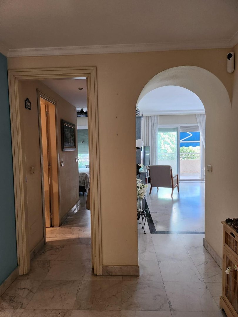 Hallway with marble floors leading to a living area with a chair and balcony overlooking a blue sky and greenery.