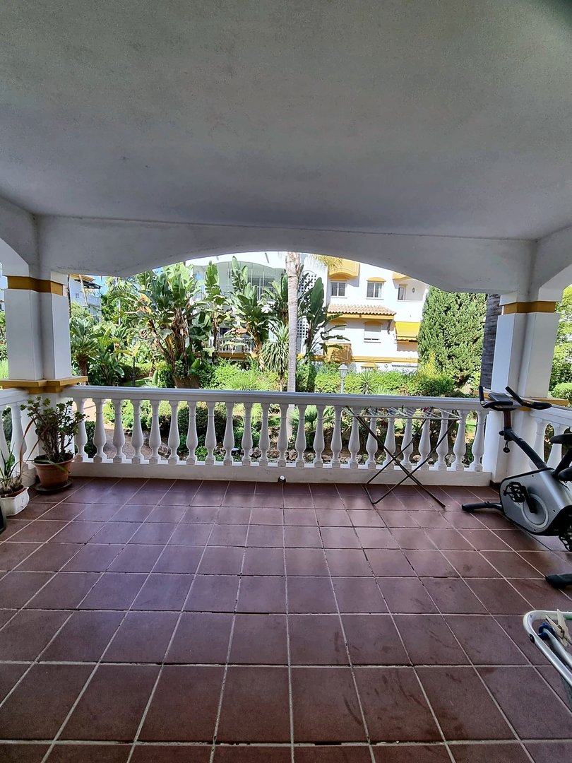 Covered terrace with white railings and potted plants overlooking a lush garden and distant buildings. Exercise equipment is visible.