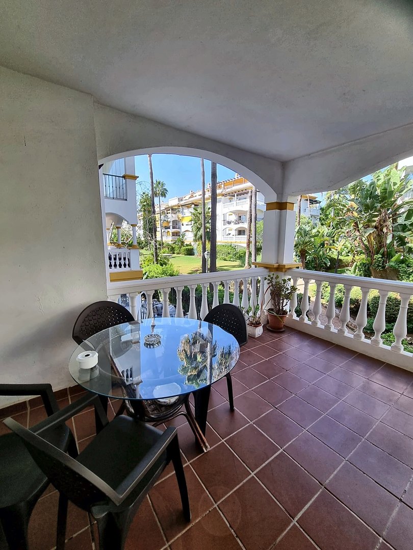 Balcony with a glass table and chairs, overlooking a resort with palm trees and buildings.