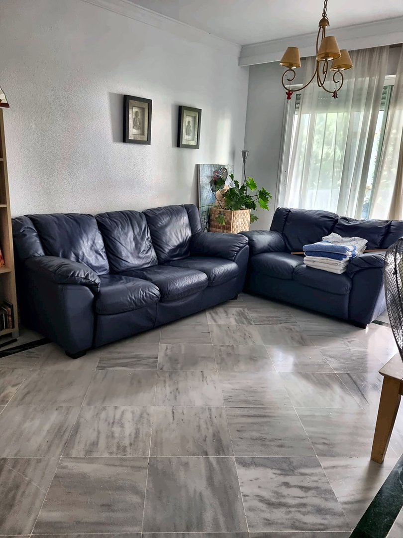 A living room featuring a dark blue leather sofa and armchair, marble flooring, and white curtains revealing natural light.