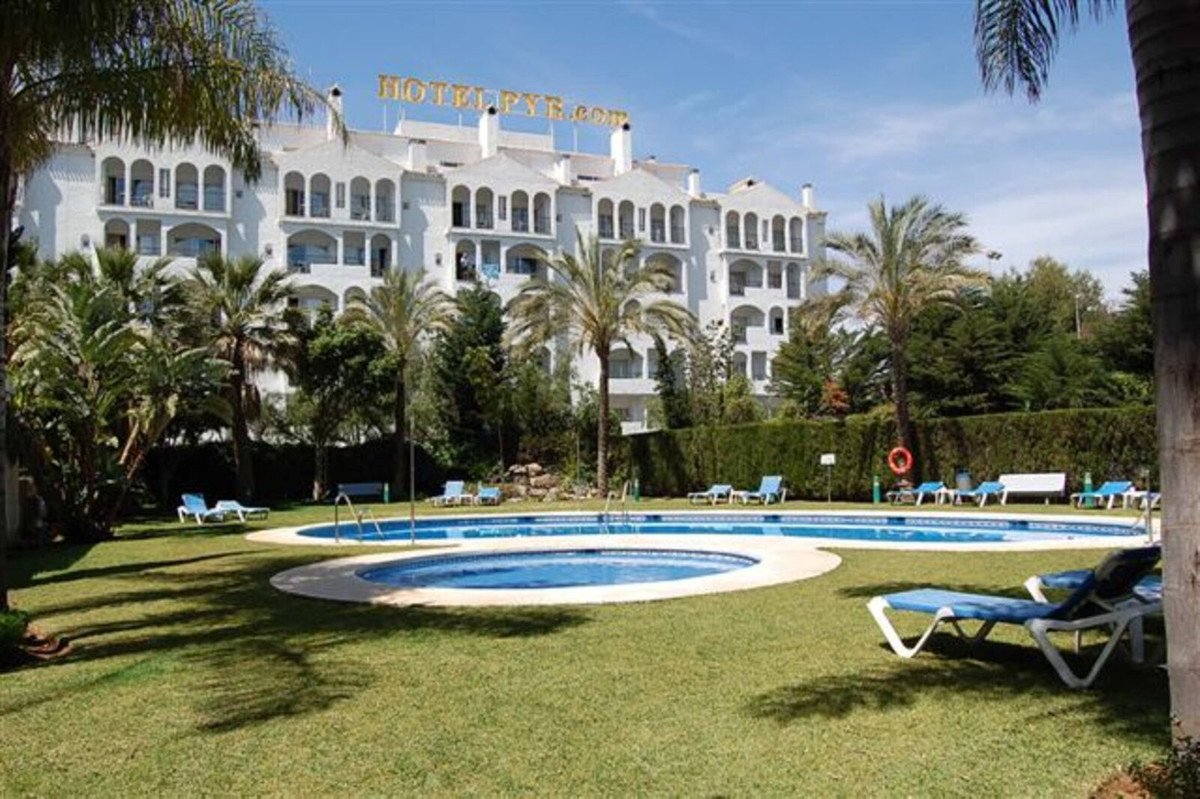 A multi-level white hotel building with a rooftop sign is surrounded by palm trees and lush greenery, featuring a swimming pool with lounge chairs.