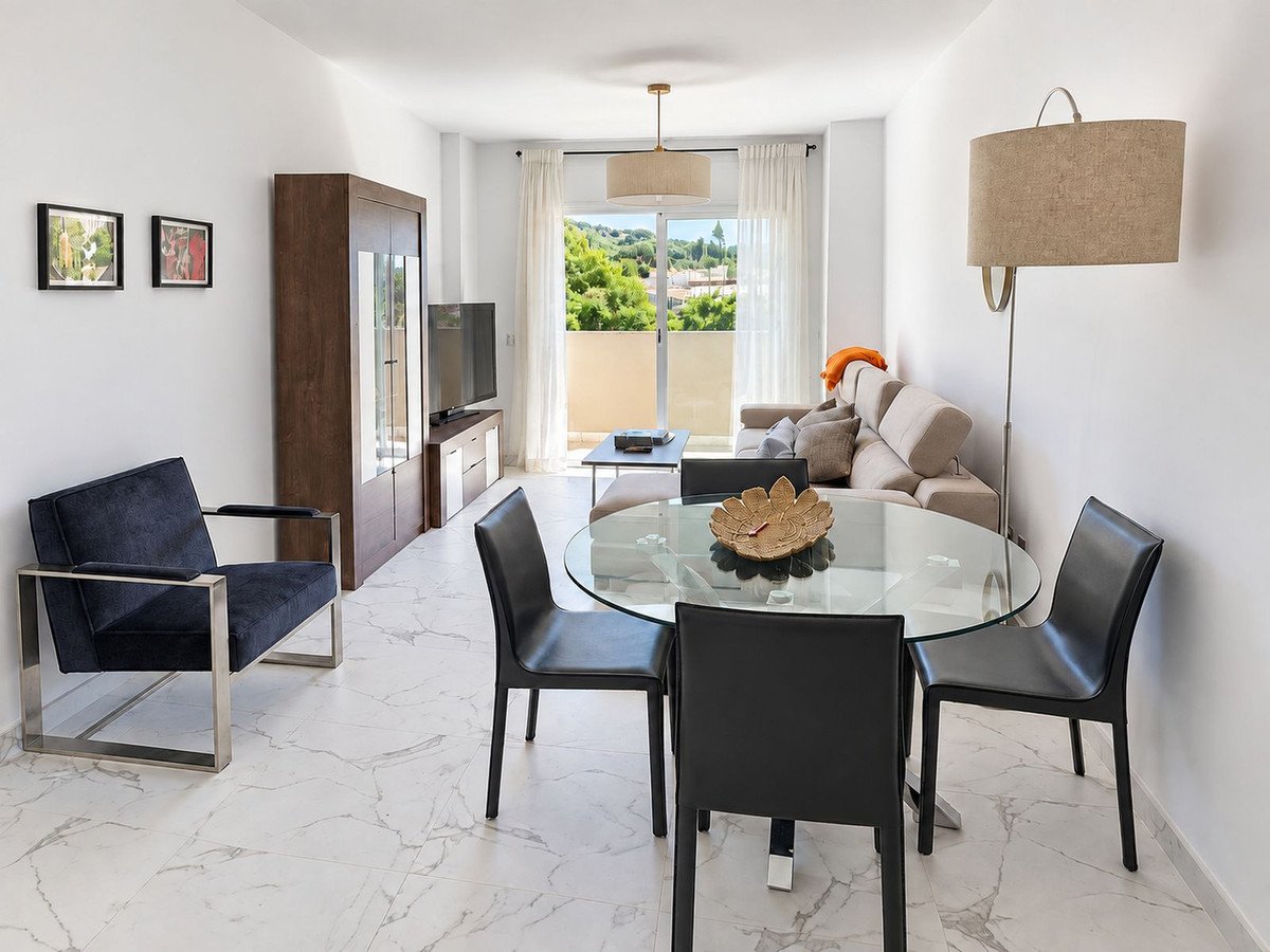 Modern living and dining area with glass table, sofa, chairs, and balcony access overlooking greenery.