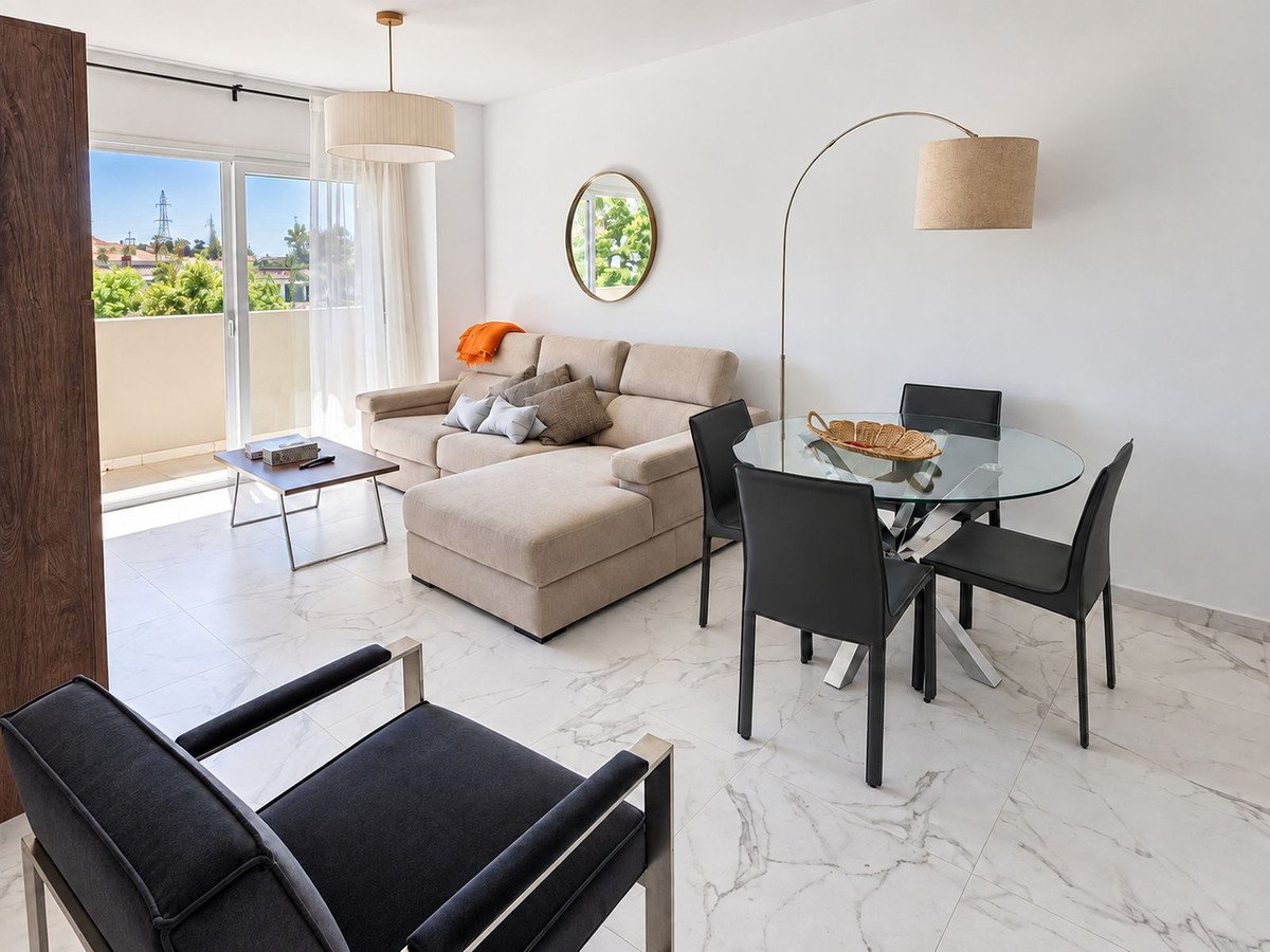 Bright living and dining area with a sectional sofa, dining table, and balcony access with a view of greenery and utility poles.