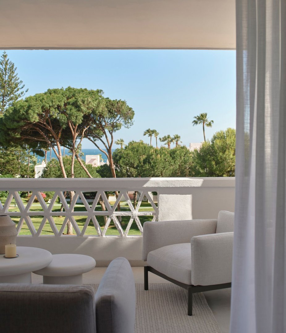 A modern balcony with comfortable seating, overlooking lush greenery and the sea under a clear blue sky.