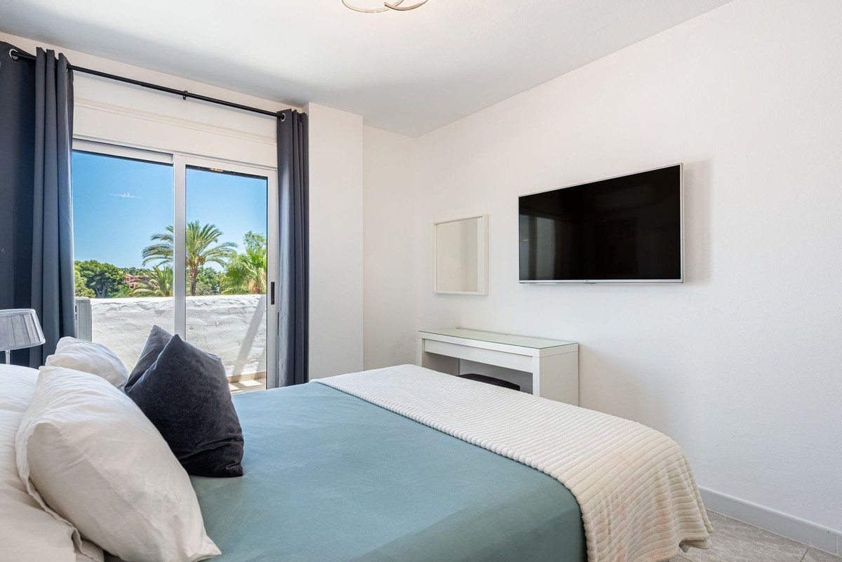 A bedroom featuring a bed, a wall-mounted TV, a dressing table, and a balcony with a view of palm trees.