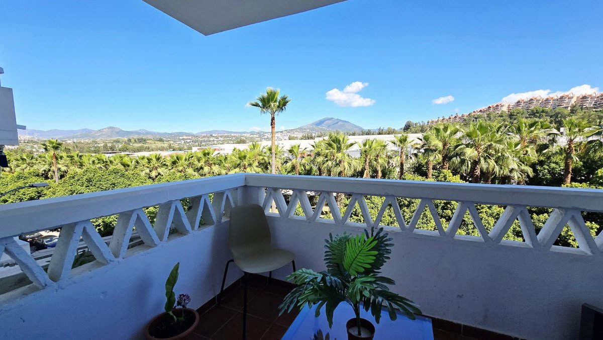 A balcony with a chair and plants offering a panoramic view of palm trees and mountains under a clear blue sky.