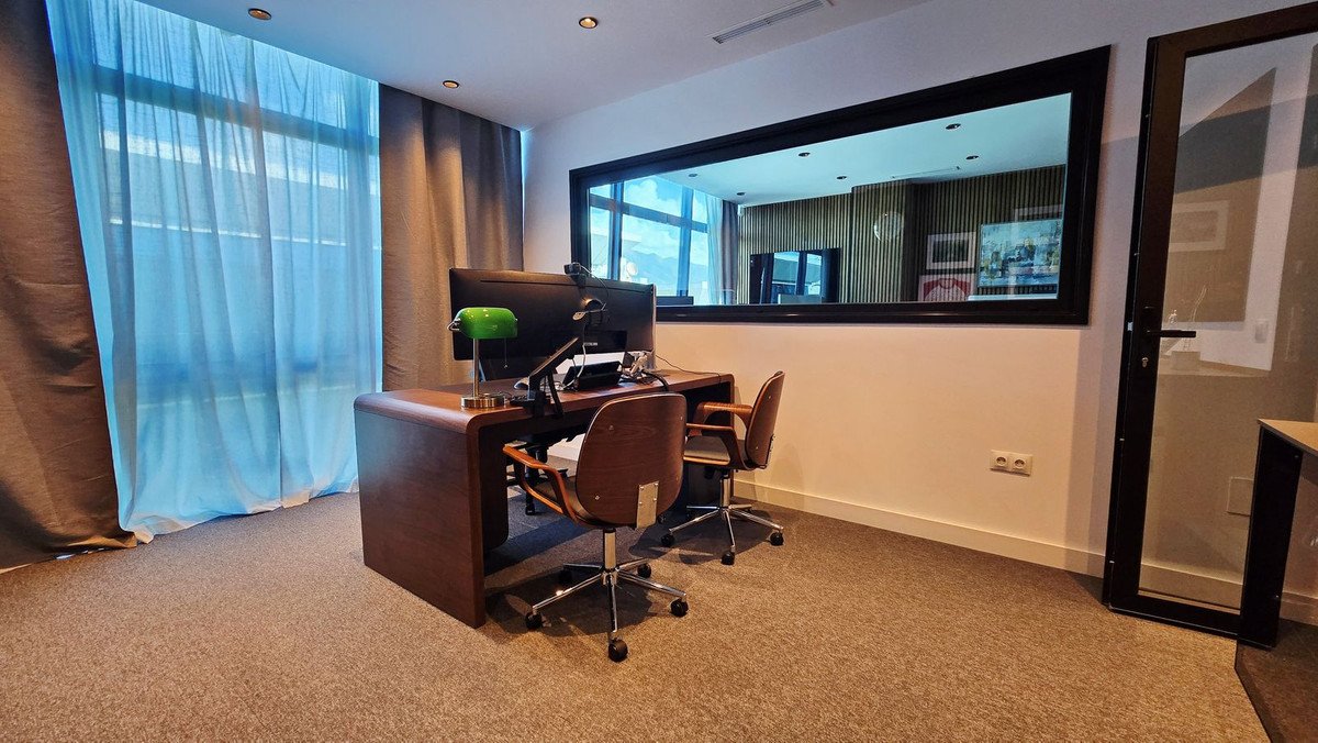 A modern office room with a wooden desk, two chairs, a large computer monitor, and sheer blue curtains offering city views.