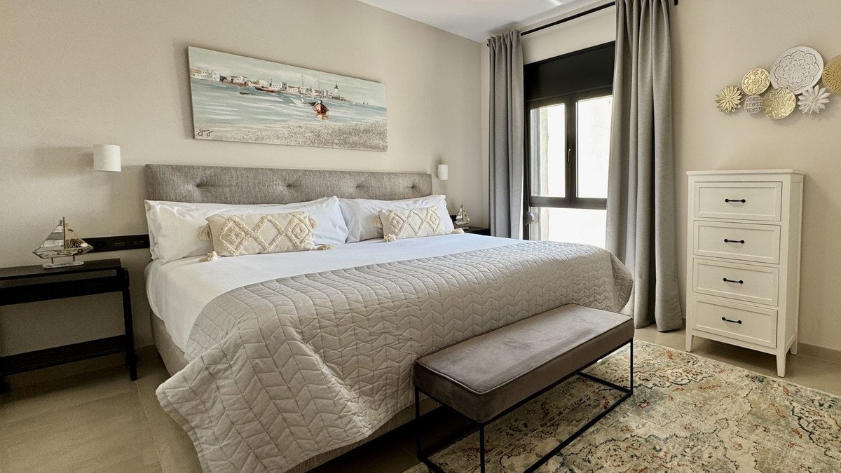 A cozy bedroom with a large bed, coastal artwork, and a window view, featuring a white dresser and patterned rug.