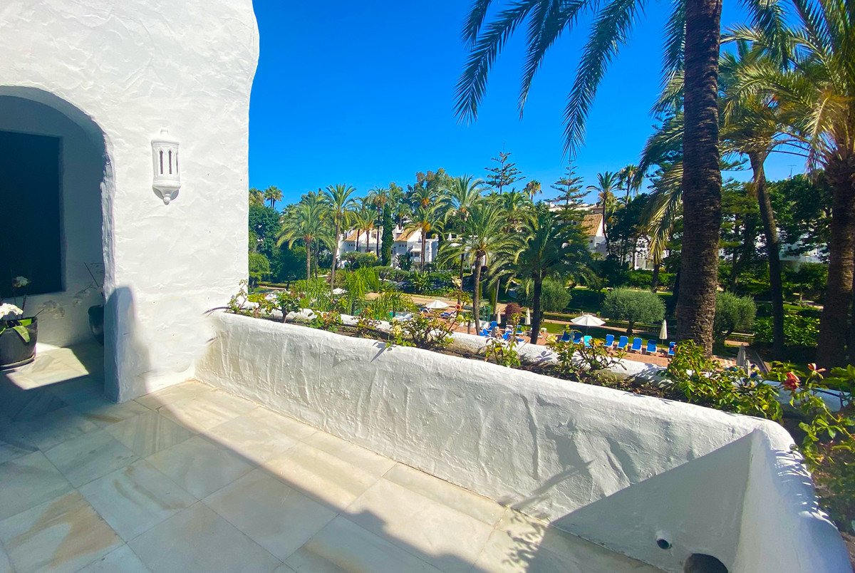 View from a white balcony overlooking a resort with palm trees, gardens, and a swimming pool on a sunny day.