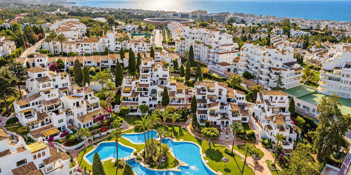 Aerial view of white apartments with pools and gardens, overlooking the sea and coastline.