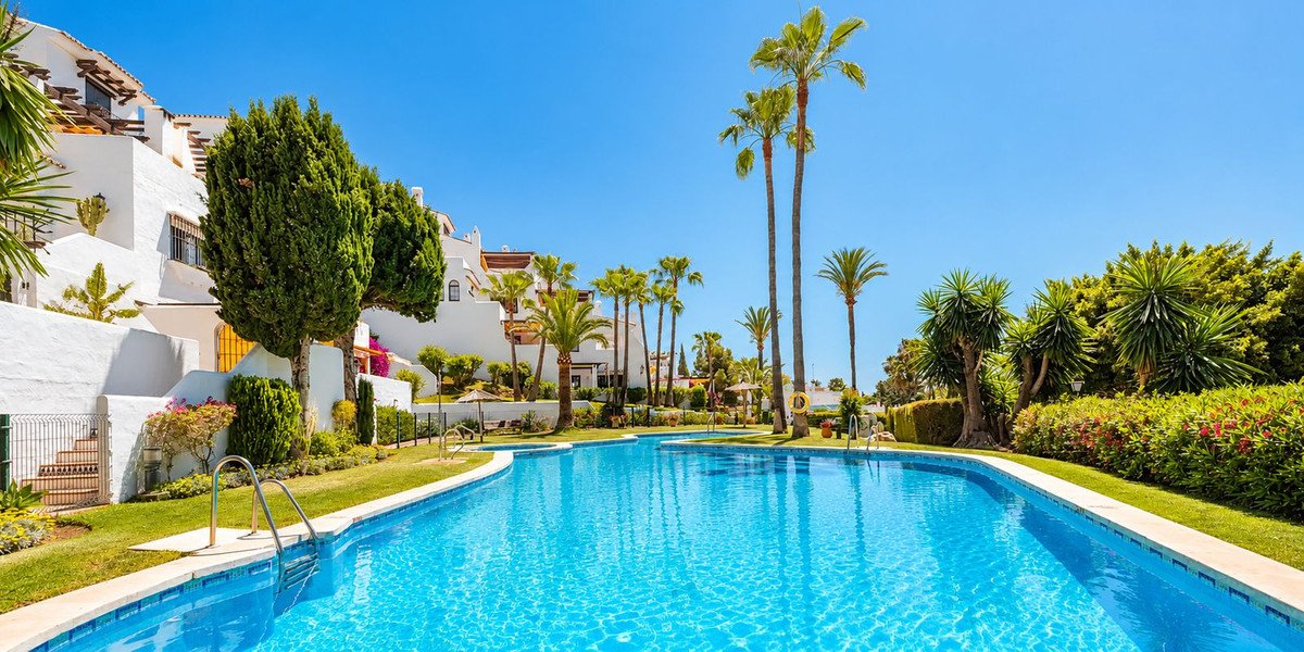 A resort-style swimming pool surrounded by palm trees and lush greenery, with white apartment buildings in the background.