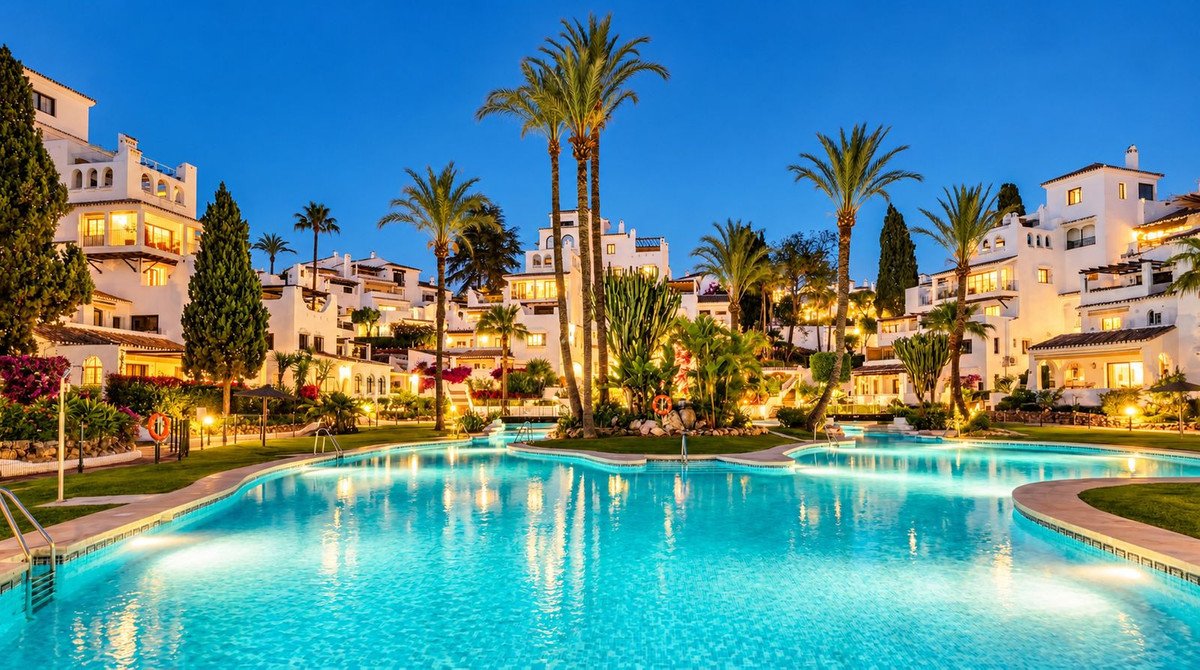 A large, illuminated swimming pool in front of white apartment buildings and palm trees at dusk.