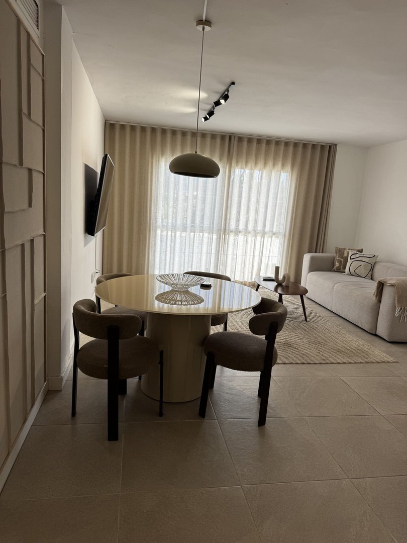 Modern dining area and living room with elegant furniture, beige curtains, and a TV mounted on the wall.