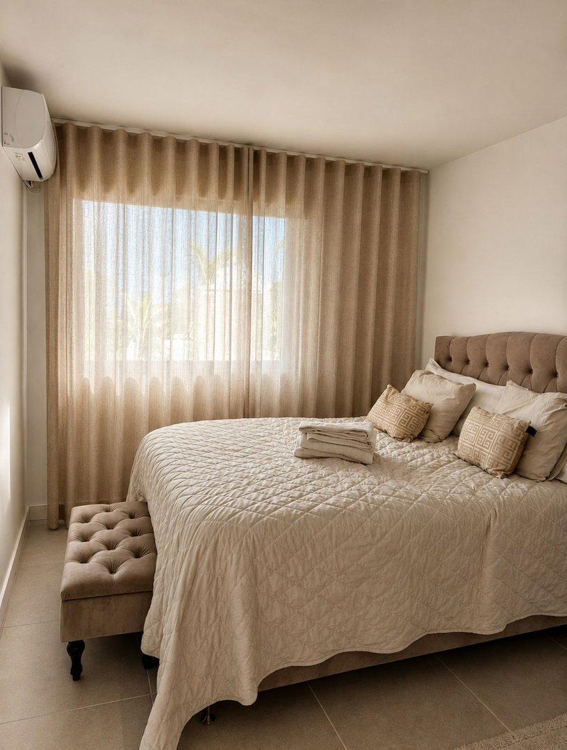 A bedroom features a large bed with a tufted headboard, plush pillows, and a quilted blanket. A decorative bench sits at the foot of the bed.