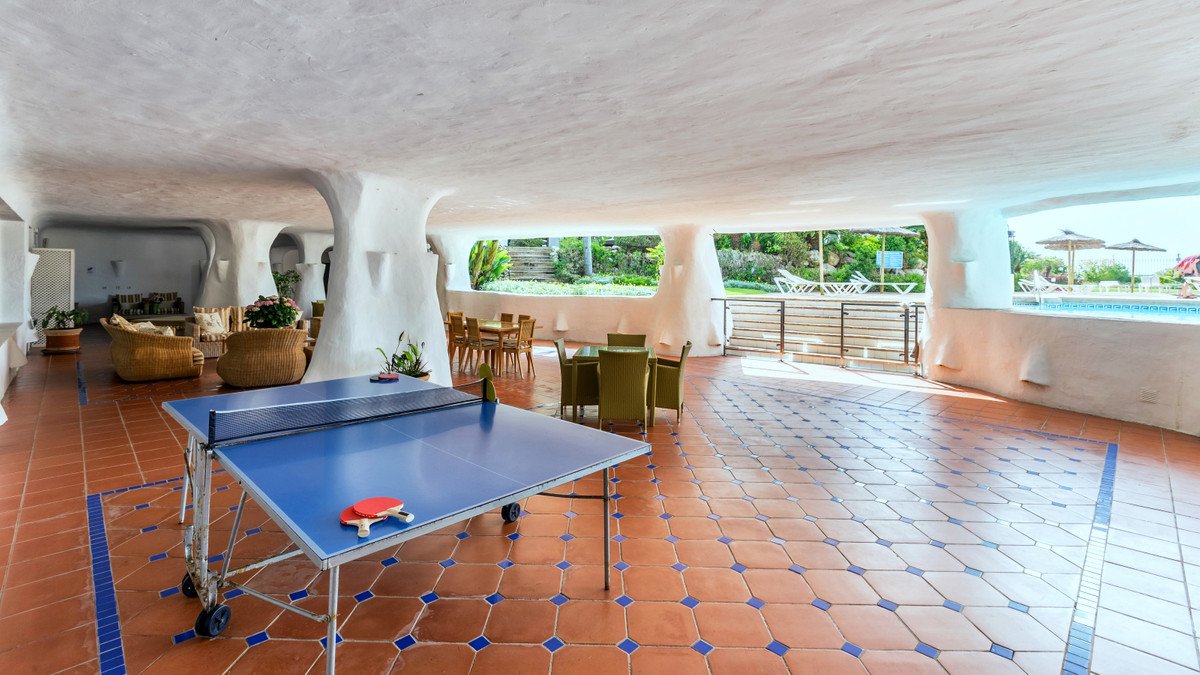 Covered outdoor area with a ping pong table, seating, dining tables, and a view of the swimming pool and gardens.