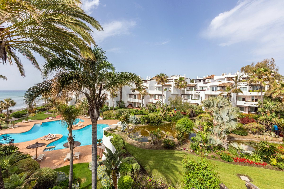 Resort complex with swimming pool, lush gardens, pond, palm trees, and ocean view in the background.