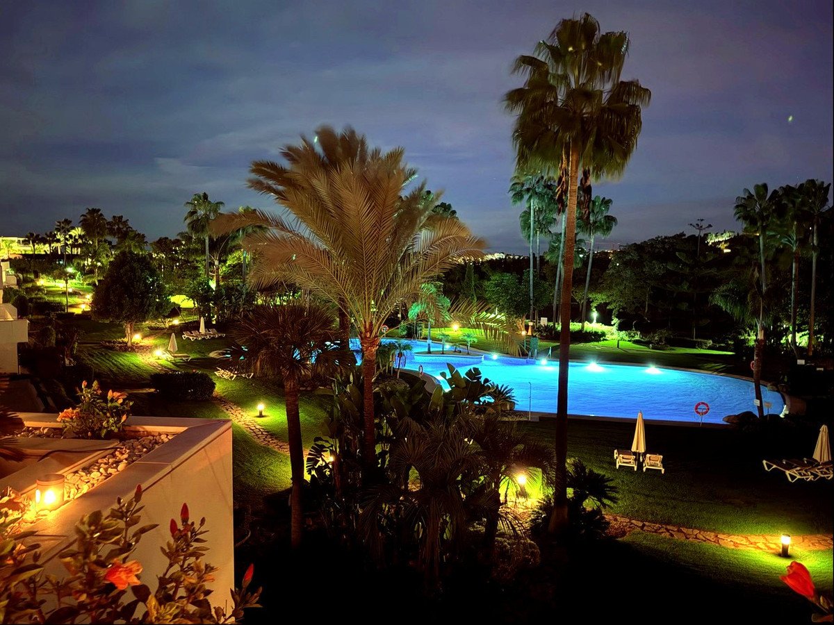 Night view from a villa, showcasing a brightly lit pool, palm trees, and lush gardens.