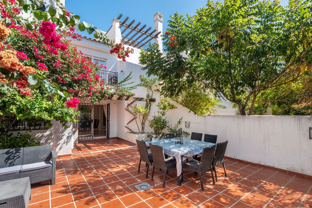 Tiled terrace with outdoor dining table, chairs, and lounge seating, surrounded by lush greenery and flowering plants.