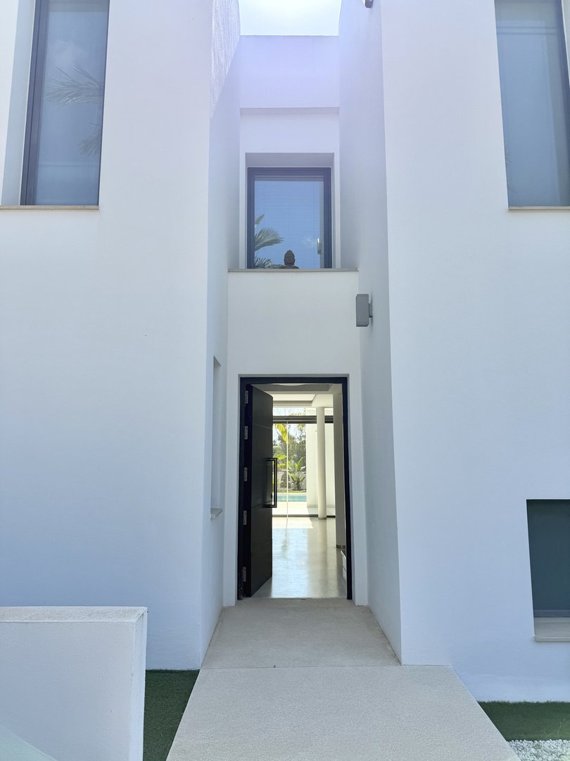 Modern white villa entrance with open door, view to pool and greenery, minimalist design.