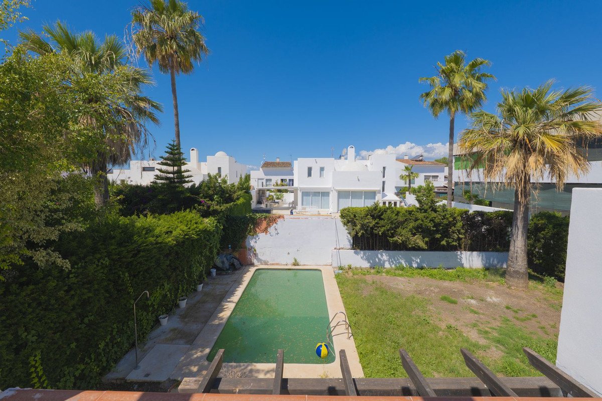 A rectangular swimming pool is surrounded by a lush green garden with mature palm trees and white houses in the background under a clear blue sky.