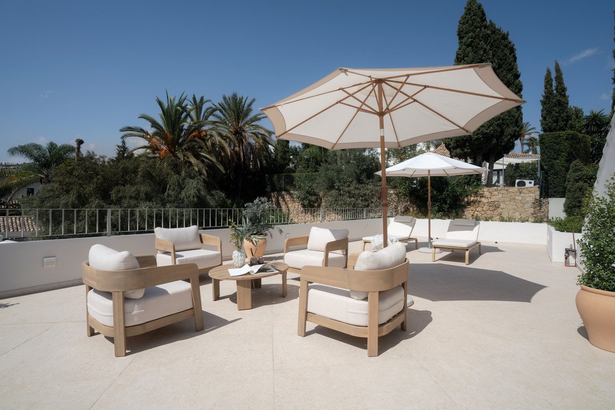 An outdoor terrace with modern white seating, wooden furniture, umbrellas, lounge chairs, and lush greenery under a clear blue sky.