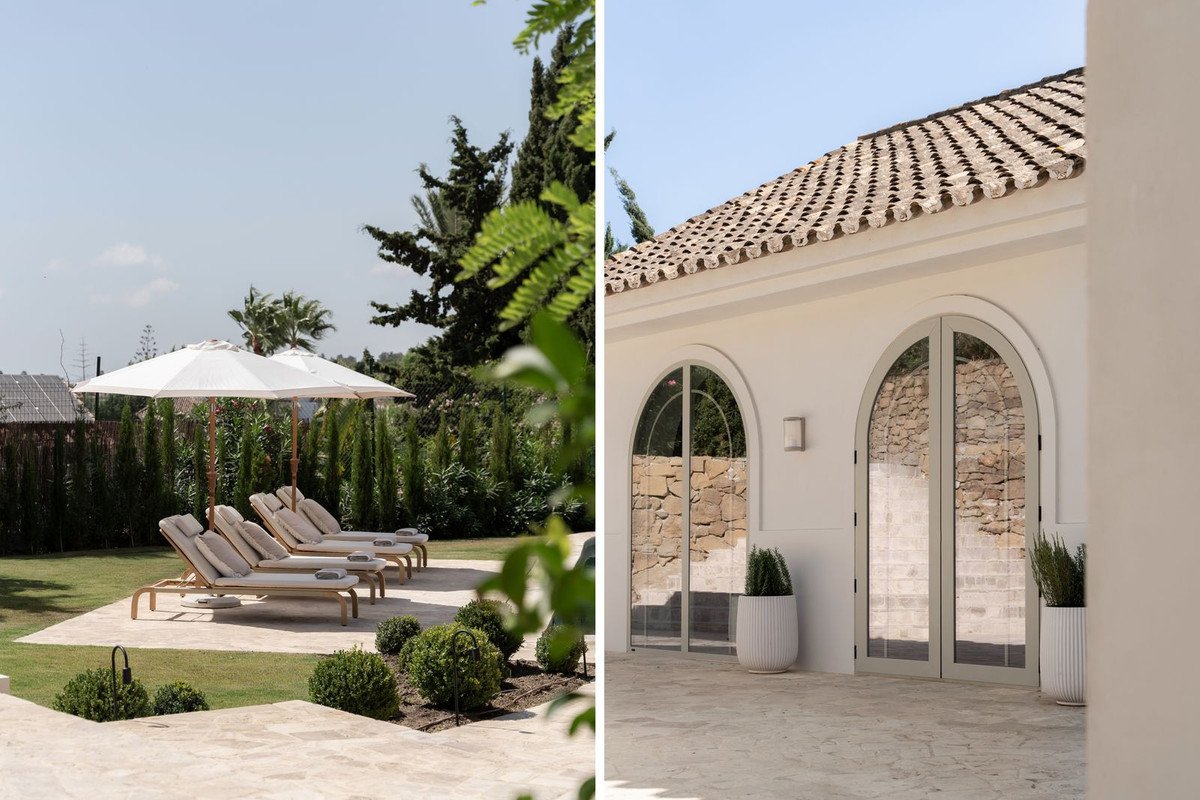 Sun loungers under umbrellas in a garden next to arched glass doors with stone walls visible inside.