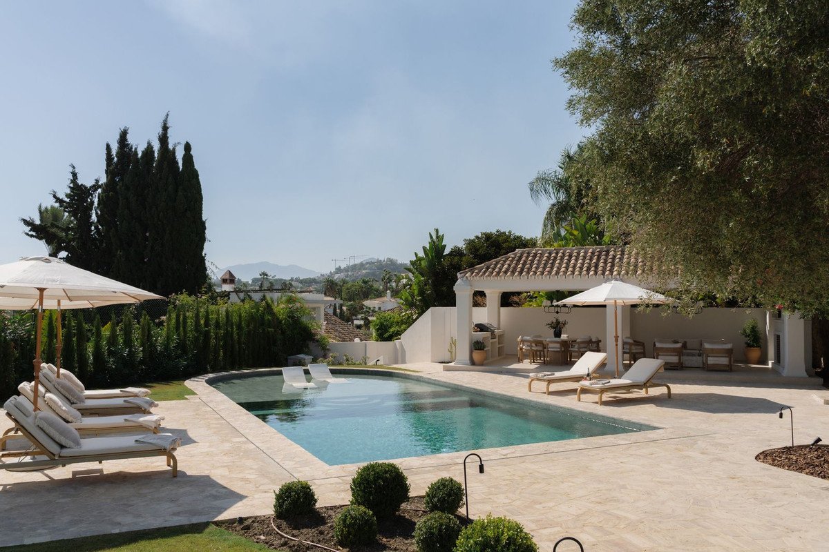 Luxurious outdoor area with a swimming pool, lounge chairs, dining table under a pergola, and a view of the hills.