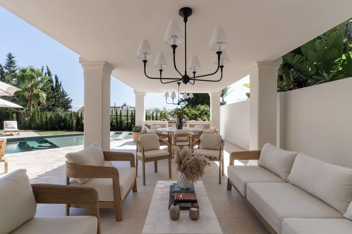 Covered outdoor patio with dining table and chairs, sofas, and lounge chairs overlooking a swimming pool and garden.