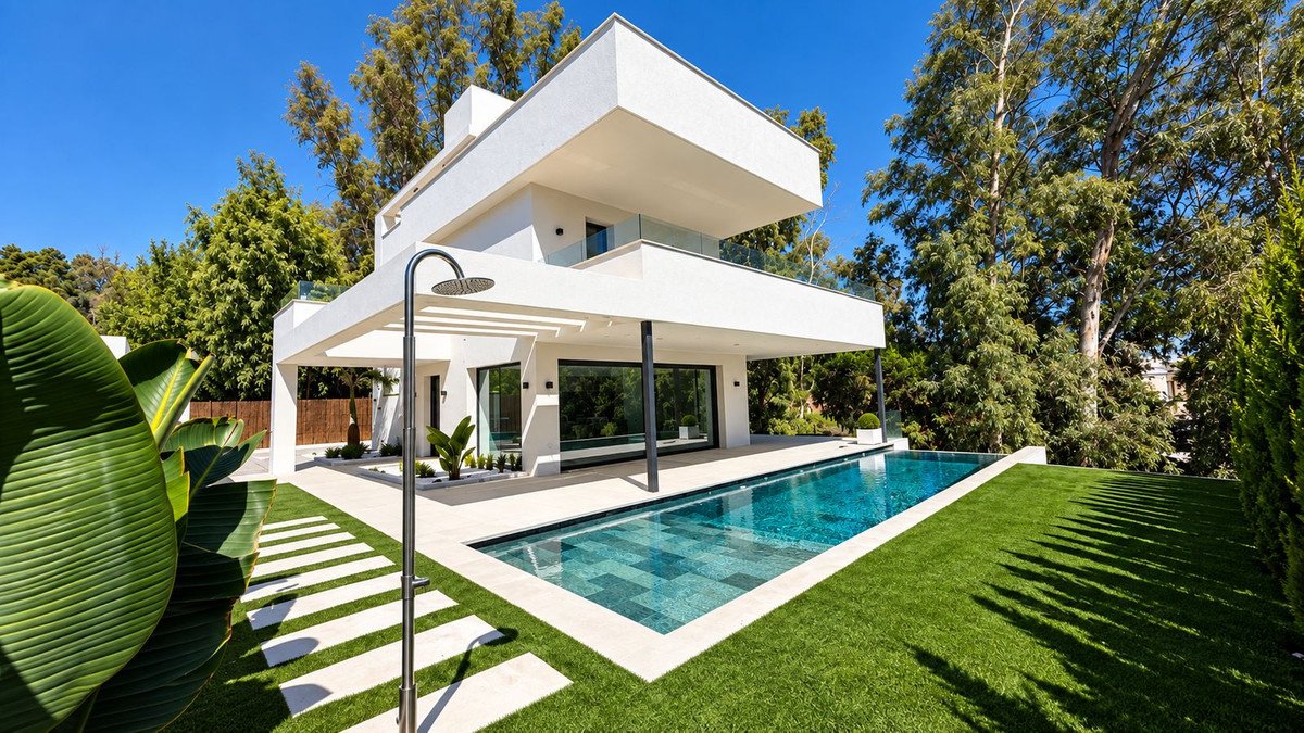 Modern white villa with a long swimming pool, outdoor shower, and lush green lawn surrounded by trees.