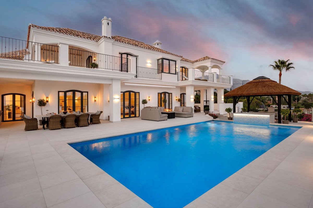Luxury villa with a large swimming pool, outdoor dining area, lounge seating, and a thatched gazebo during sunset.