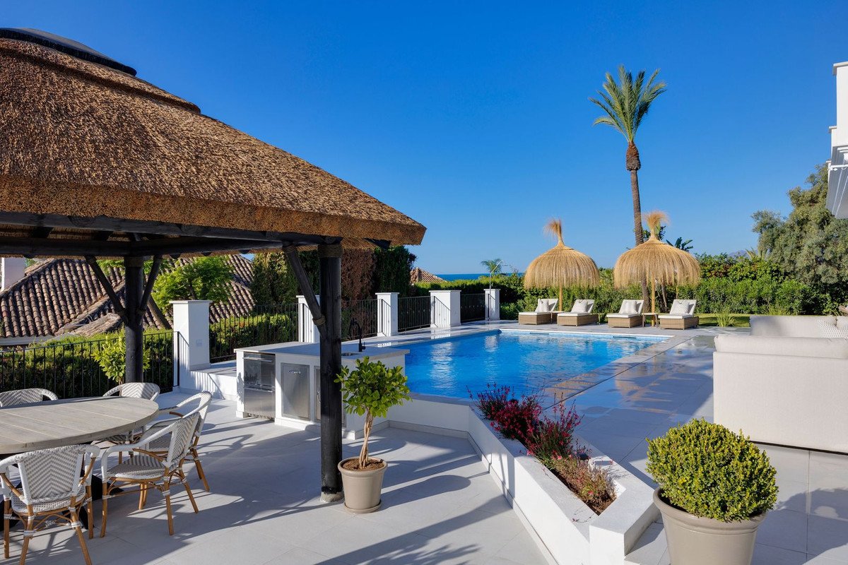 Luxury outdoor area with a swimming pool, sun loungers, dining table, and a thatched-roof cabana, overlooking the sea.