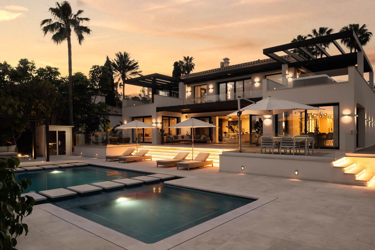A modern villa with a swimming pool and lounge chairs at sunset, featuring outdoor dining and palm trees.