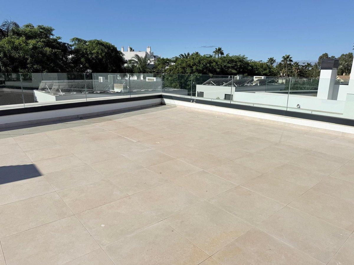 A spacious terrace with light beige tiles and a glass balcony railing, offering views of greenery and modern buildings under a clear blue sky.