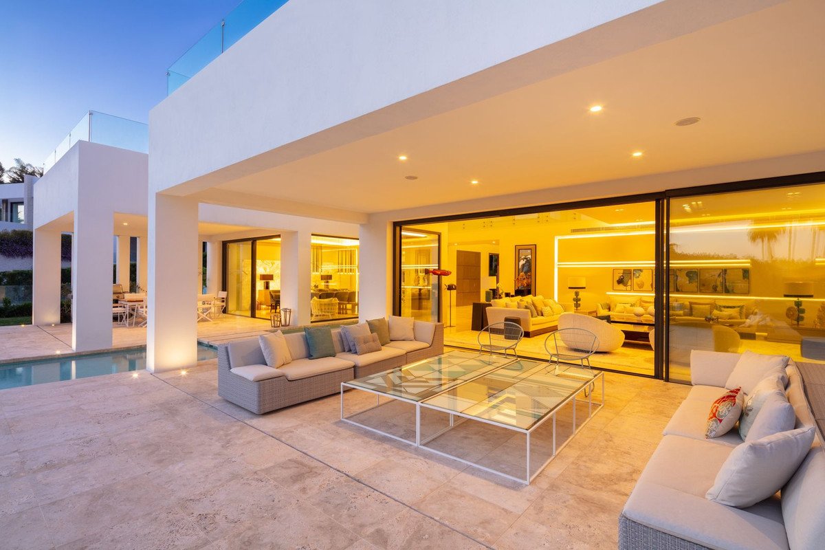 Modern luxury villa patio with comfortable seating, a glass coffee table, and a swimming pool. Large sliding doors open to a brightly lit living area.