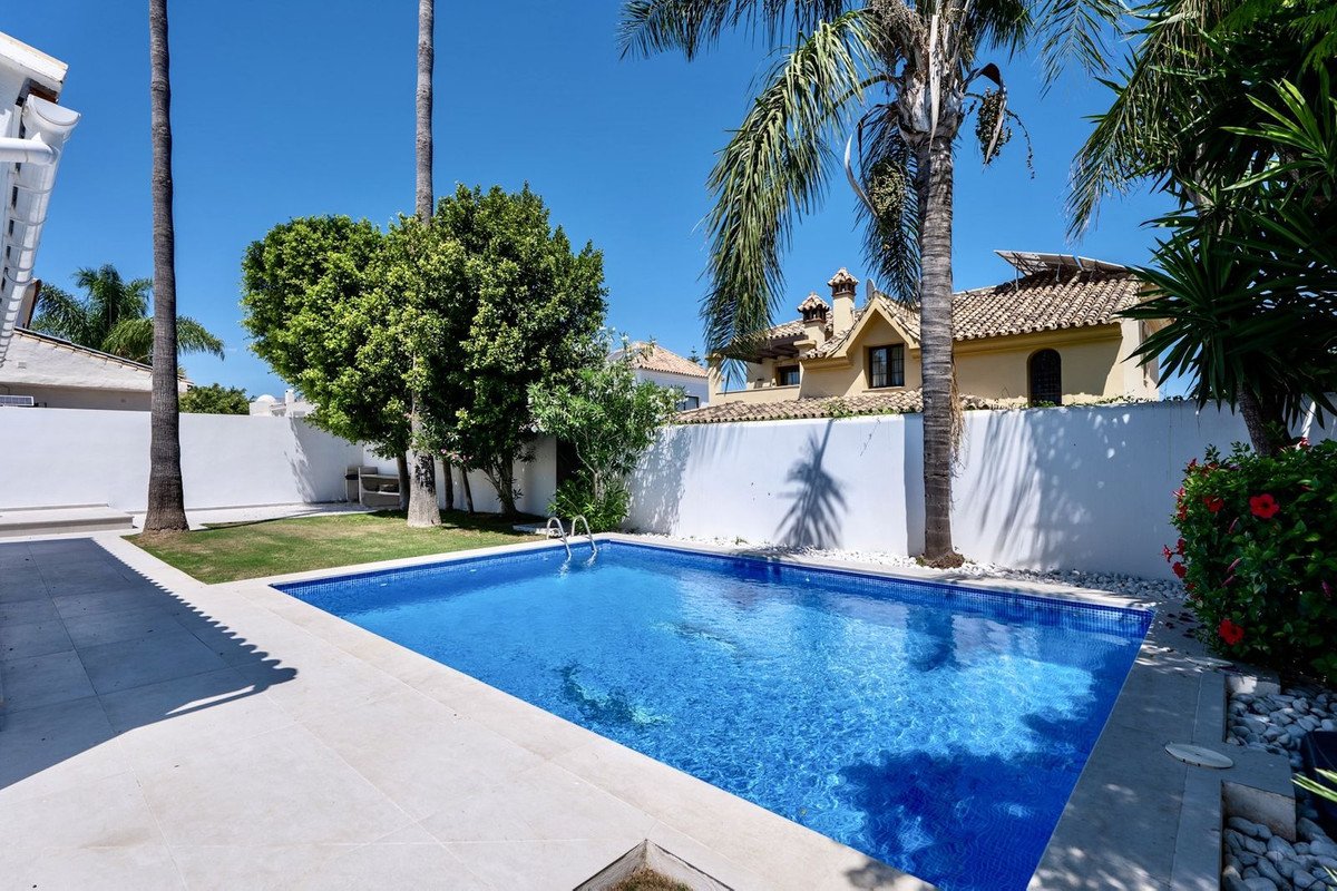 A bright blue swimming pool surrounded by a paved area and green lawn, with palm trees and a villa in the background.