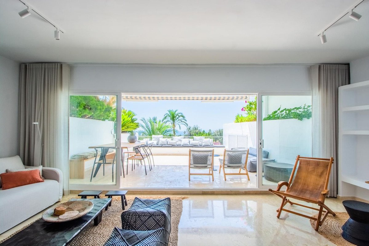 Modern living room with large sliding doors opening to a terrace with outdoor seating, overlooking the sea and palm trees.
