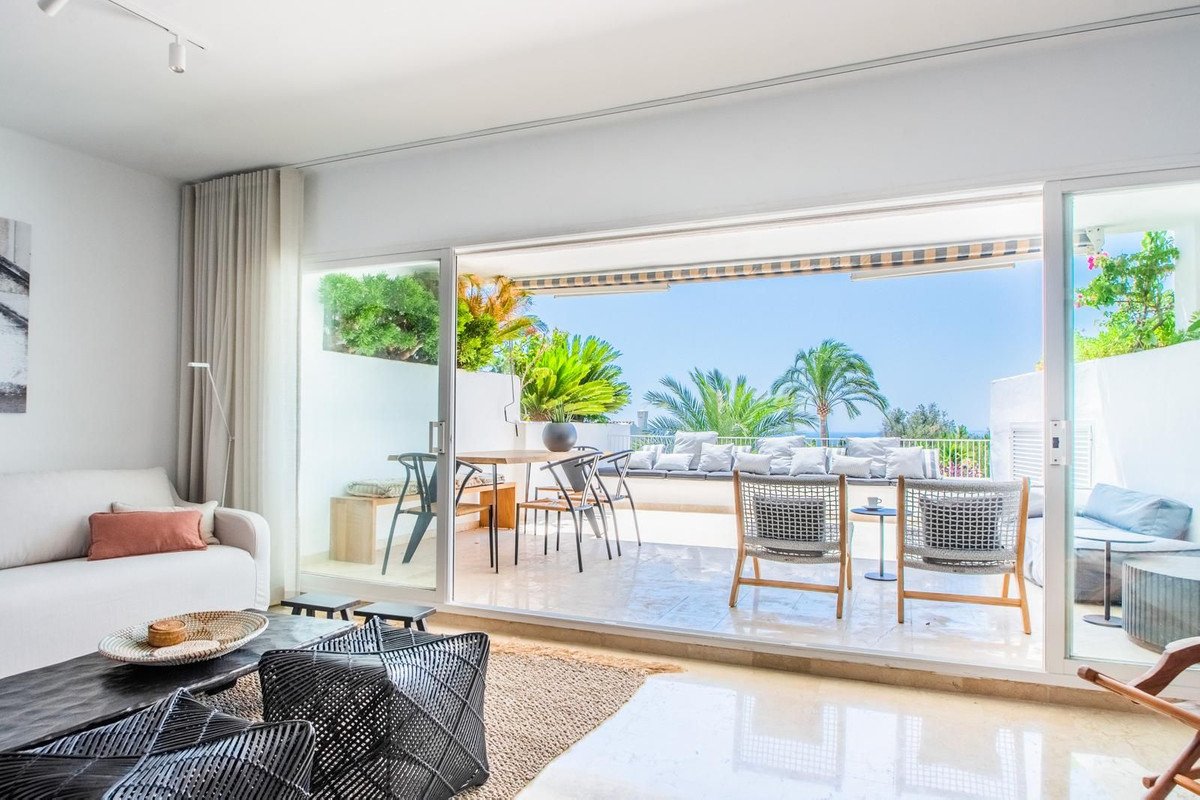 Spacious living room opening onto a terrace with dining area, lounge seating, and a beautiful sea view under palm trees.
