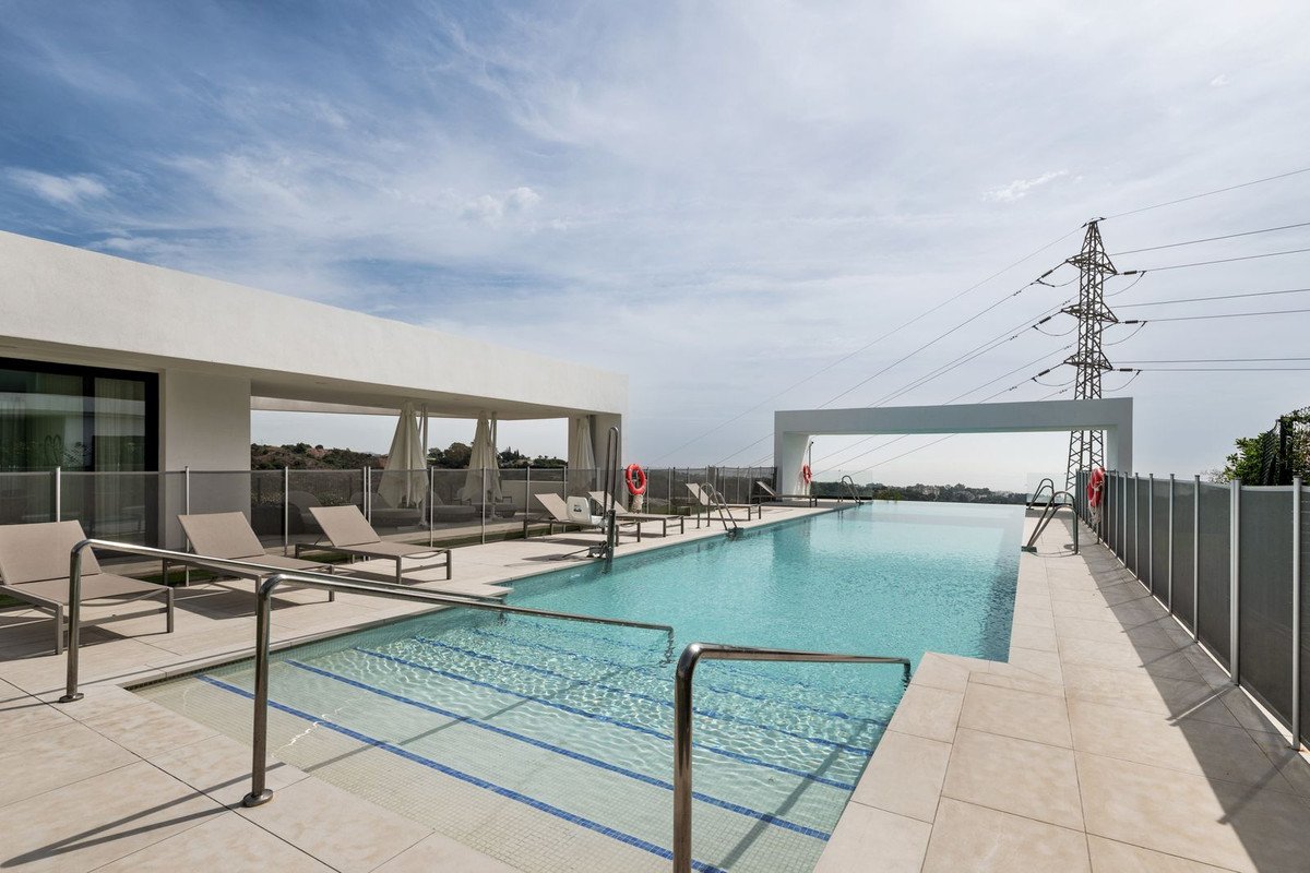 Infinity swimming pool with lounge chairs on a rooftop terrace under a cloudy blue sky with distant hills and a power line tower.
