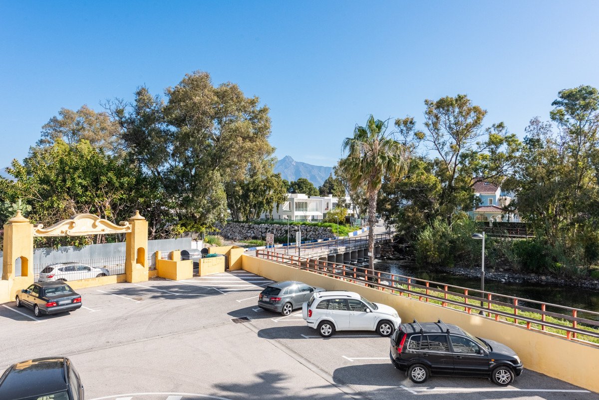 Parking area with several cars, a yellow arched entrance, and a distant view of mountains and modern houses.