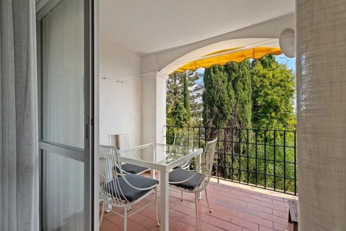 A small balcony with a table and four chairs, featuring a view of lush green trees and blue sky, with a retractable awning overhead.