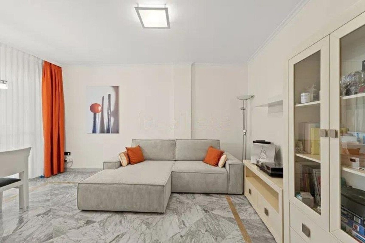 A modern living room with a grey sectional sofa, orange accent pillows, marble floors, and bright orange curtains.