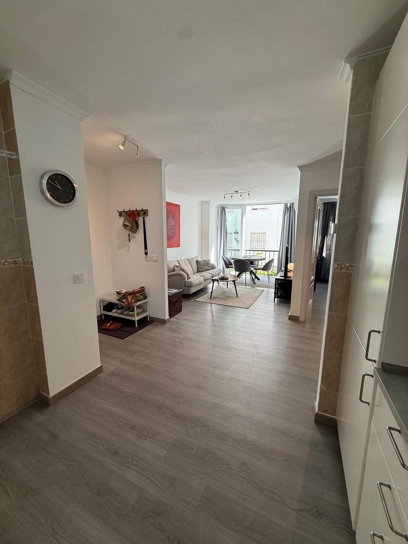 A bright living room with a sofa, coffee table, and a view of the balcony and dining area through the open doorway.