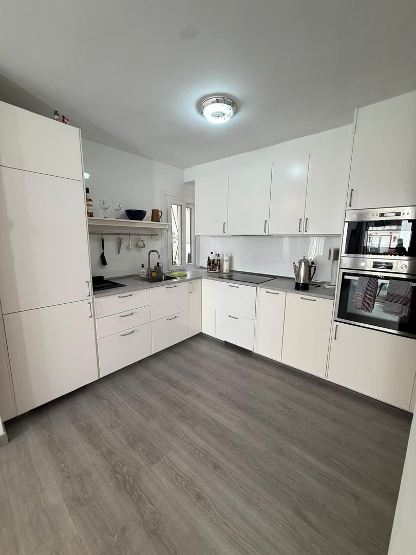 Modern white kitchen with gray wood-look flooring, white cabinets, integrated appliances, and a view through a window.