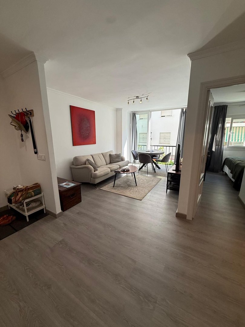 A bright living room with a sofa, coffee table, and dining area. Large windows lead to a balcony with a city view.
