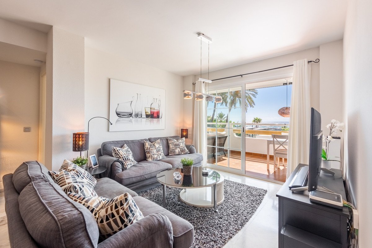 Modern living room with grey sofas, coffee table, and sliding glass doors opening to a balcony with sea and palm tree views.