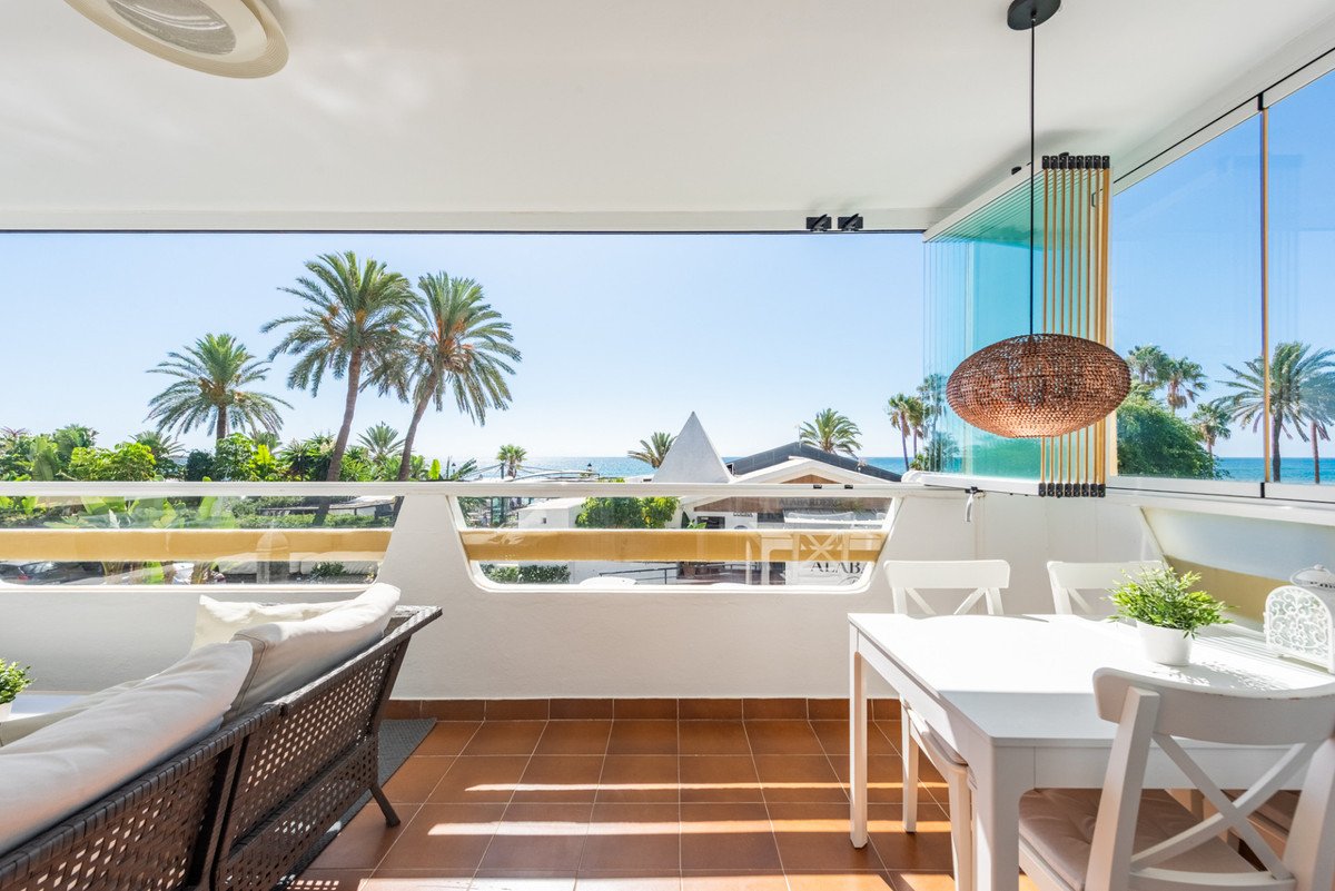 Bright balcony with wicker sofa, dining table, and sea view with palm trees.