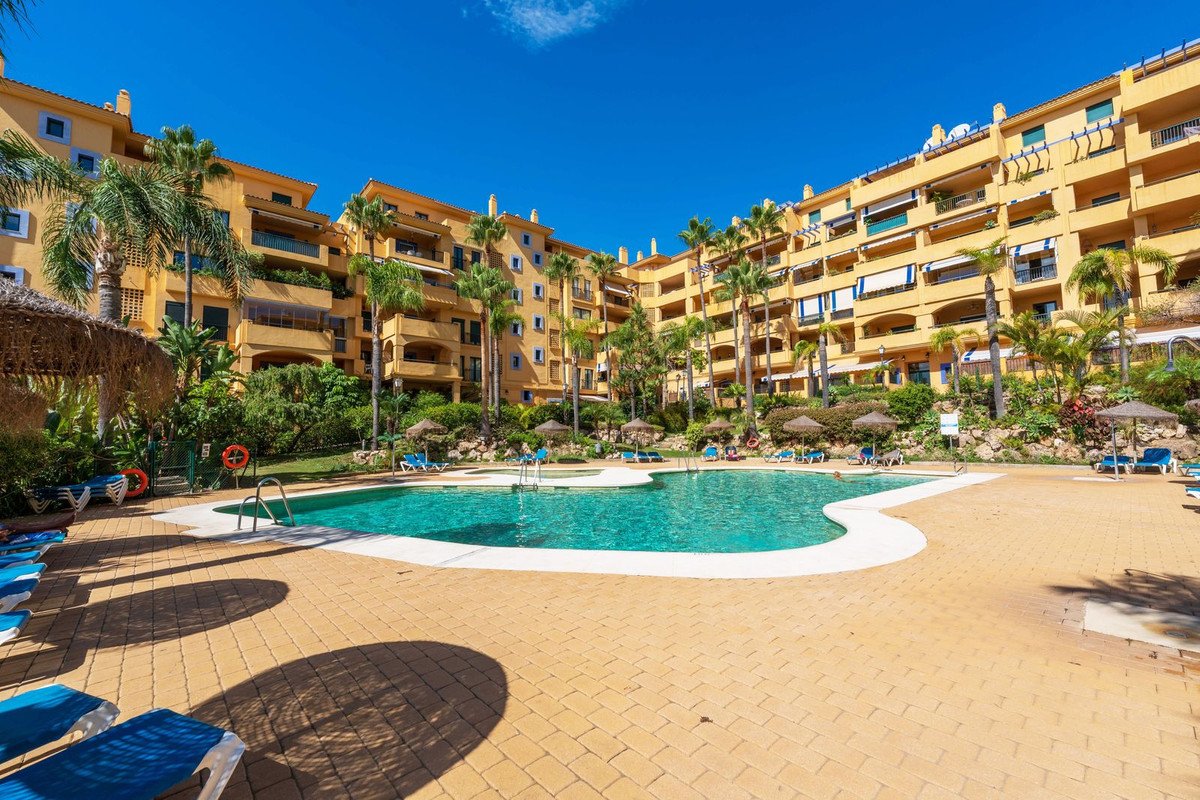 A large, resort-style swimming pool with sun loungers and palm trees in front of a yellow apartment building under a blue sky.