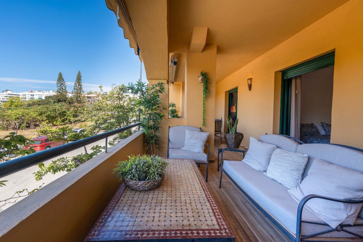 A balcony with outdoor seating and a table, overlooking greenery and buildings under a clear blue sky.