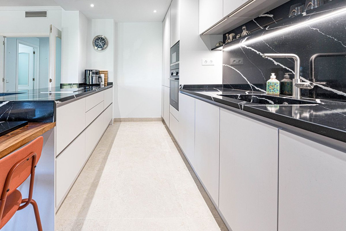 Modern kitchen with white cabinets, black marble countertops, a sink, faucet, and built-in oven. Illuminated backsplash.